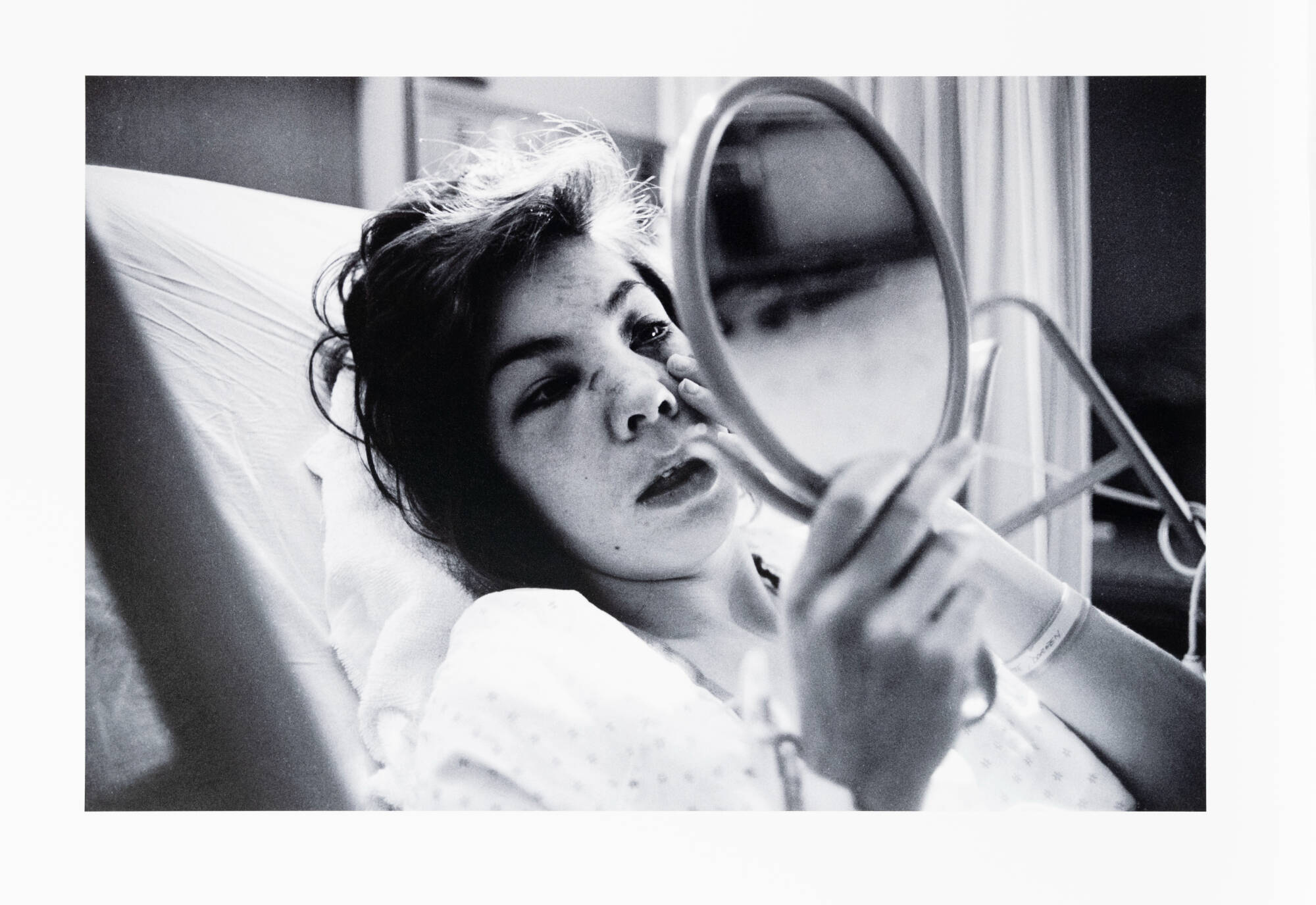 Black and white photograph of a woman in a hospital bed, looking in a mirror.